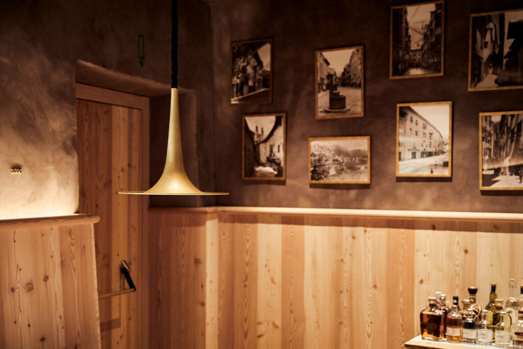 Ein mit Holz getäfelter Innenbereich des Wirtshaus Decantei in Brixen. Zentral ist eine trompetenförmige Messinglampe, im Hintergrund sieht man historische schwarz-weiß Fotos der Stadt Brixen.