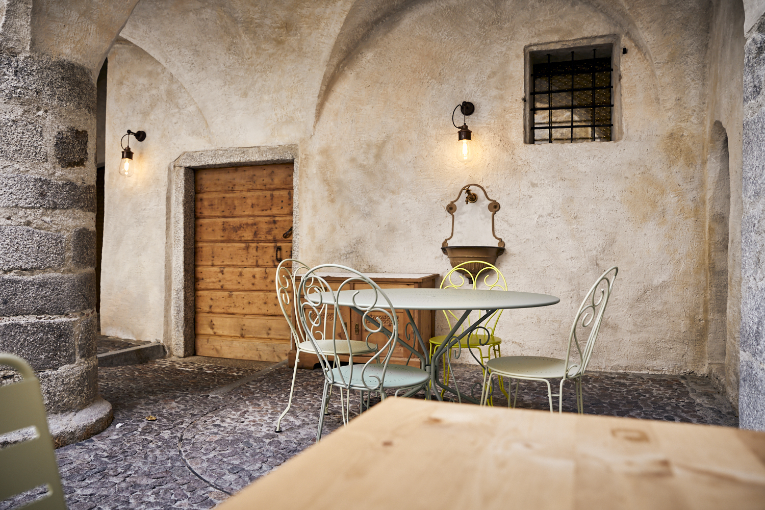 Ein türkis-grüner Tisch im Innenhof des Wirtshaus Decantei in Brixen. Im Hintergrund sieht man eine rustikale Mauer, ein Holztor und ein zwei brennende Lampen.